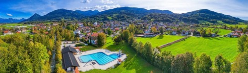 Panoramic view of a village with a swimming pool, surrounded by green meadows and forests, against