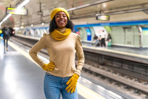 Smiling young woman wearing yellow wool cap and gloves is waiting for subway train arrival at