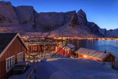 Red wooden houses on a fjord in front of snow-covered mountains, illuminated, twilight, winter,