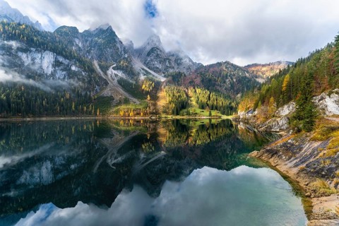 The Vordere Gosausee in autumn. The Gosaukamm on the left. Clouds and sun. Trees in autumn leaves.