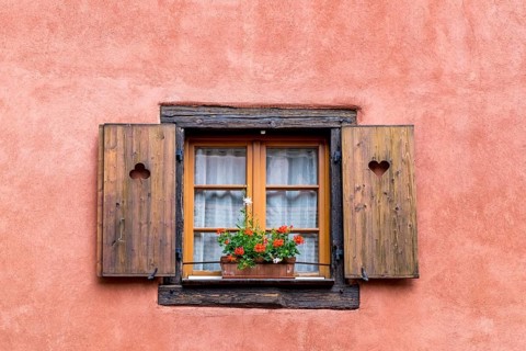 Old window with wooden shutters and flower box, framed by a pink wall, creates a rustic and