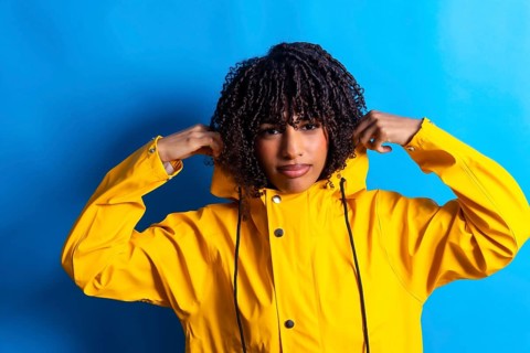 Studio shot of a young woman with curly hair adjusting the hood of her yellow raincoat against a