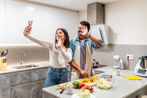 Young couple waving at the camera while preparing a healthy salad in their kitchen, making a video
