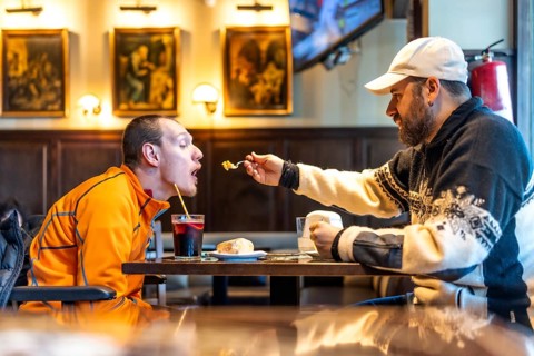 Man feeding his disabled friend in a restaurant, showcasing genuine care and inclusivity while