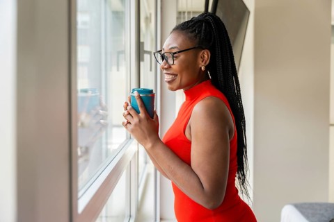 Smiling businesswoman drinking from a reusable cup while looking out the window of her hotel room