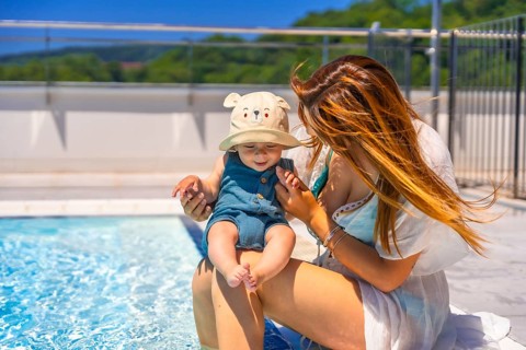 Adorable baby with funny hay and mother sitting on the edge of a pool in a sunny day of summer