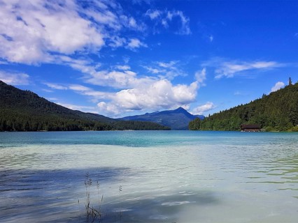 A clear lake with surrounding mountains and wooded slopes under a blue sky with white clouds,