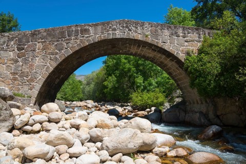 An old stone bridge spans a shallow river, surrounded by stones and trees under a clear sunny sky,