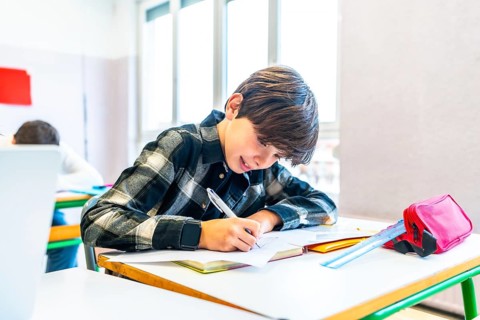 Young boy writing with pen on paper at desk in school classroom, smiling and focused during lesson