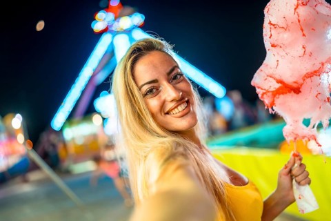 Woman taking selfie while eating pink cotton candy at fair during summer night