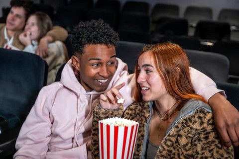 Happy diverse couple enjoying film and sharing popcorn bucket in movie theater, whispering during