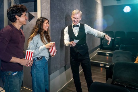Young couple holding popcorn buckets while usher showing their seats in a movie theater, ready to