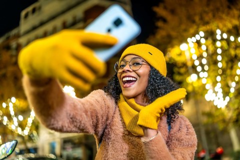 Smiling woman wearing yellow woolen gloves, hat and scarf taking selfie with her smartphone in a