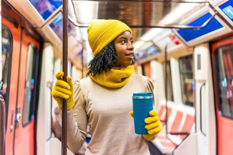 Black woman wearing yellow winter clothes holding a blue reusable coffee cup standing on a subway