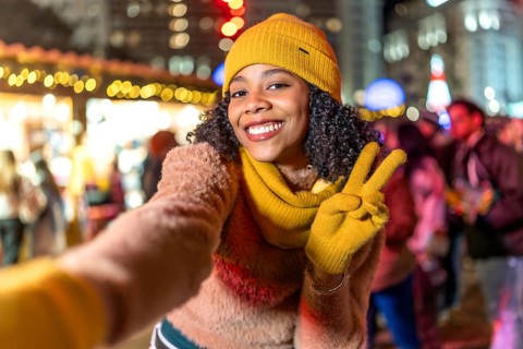 Smiling young woman wearing yellow winter clothes takes a selfie at christmas market showing peace