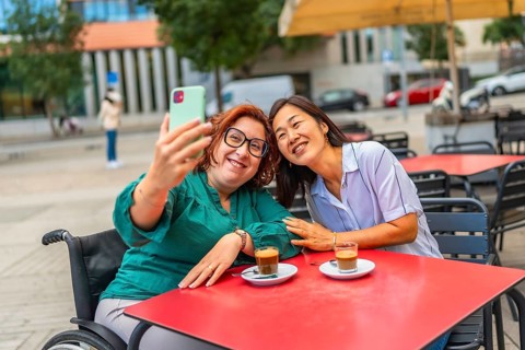 Woman with disability and asian female friend taking selfie in an outdoor cafeteria