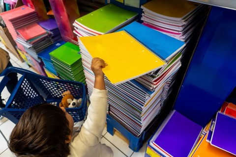 A child is reaching for a yellow spiral notebook in a store. The store is filled with many