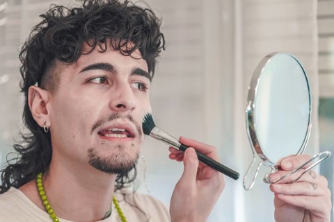 Young man with curly hair and piercings is applying makeup to his face using a brush while looking