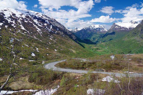 A winding road runs through a green valley between snow-capped mountains under a blue sky with