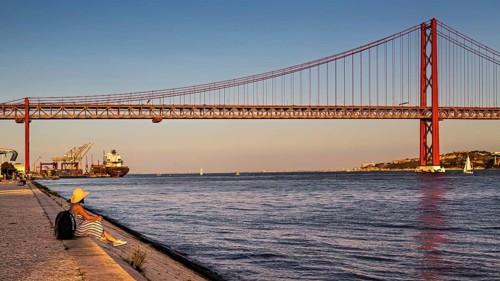 A person sits by a riverbank, gazing at the bridge and boats during a calm sunset, 25 de Abril