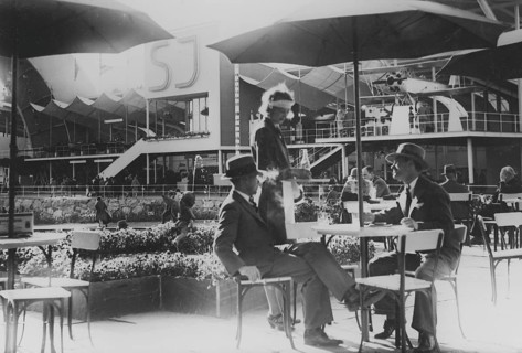 1930s open-air cafe. Two men are being served drinks at the table by a waitress on the grounds of
