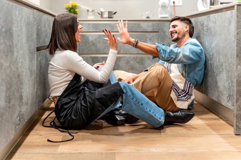 Two young chefs are sitting on the kitchen floor, giving each other a high five and laughing,