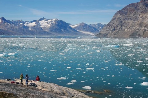 People sitting on the shore with a view of a vast icy landscape and majestic mountains under a blue