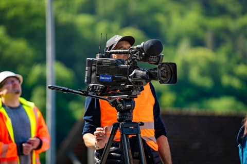 A cameraman in a brightly coloured waistcoat with a camera films outside in sunny weather against a