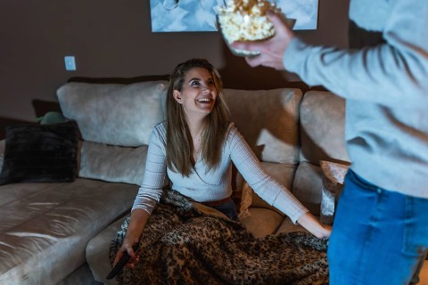 Smiling woman sitting on sofa with remote control, covered with blanket, taking popcorn from
