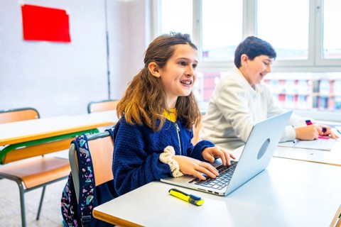 Smiling elementary school girl using laptop during lesson in classroom, next to classmate writing