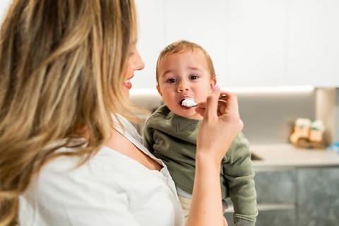 Smiling mother feeding yogurt to her baby son with a spoon in a bright kitchen, healthy eating