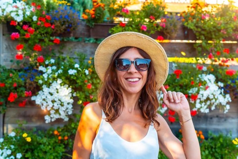 A woman wearing a straw hat and sunglasses is smiling in front of a wall of flowers. The flowers