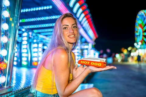 Blonde skinny woman eating a hot dog in an amusement park at night