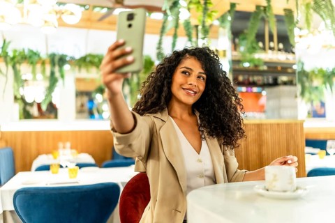 Happy and beauty latin woman with long curly hair and chic clothes taking selfie sitting alone in