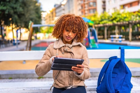 Latin beauty female student using digital tablet sitting on an urban park bench in the city