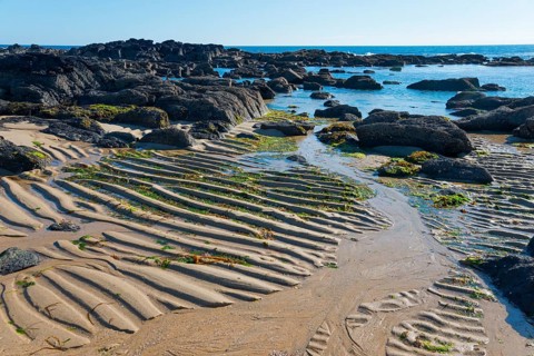 Rocks and sea with sandy wave patterns on the shore under a clear blue sky, Montedor, Viana do