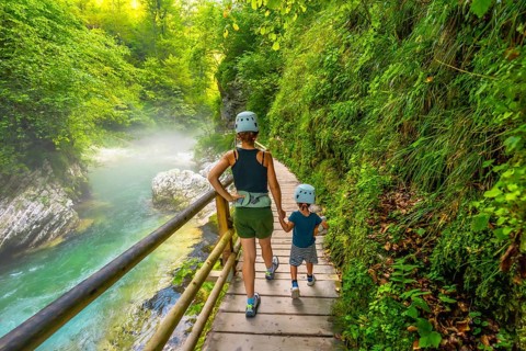 Mother and son wearing helmets are walking on a wooden pathway alongside a pristine river, enjoying