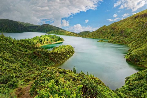 A tranquil lake surrounded by green mountains and wooded slopes under a blue sky, crater lake Lagoa