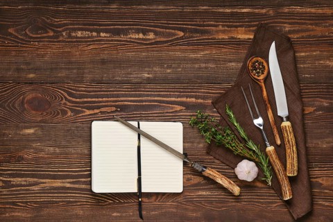 Vintage steak fork, knife and sharpener on wooden table next to notebook. Barbecue appliances and