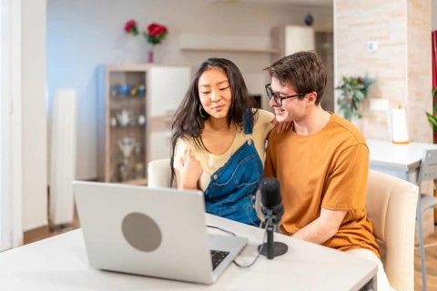 Multiethnic young couple using a laptop and a microphone, recording a podcast from their home