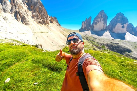 Hiker taking selfie and making thumbs up gesture with tre cime di lavaredo in background,