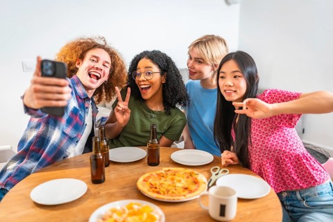 Four diverse friends are taking a selfie while enjoying pizza and beer, celebrating friendship and