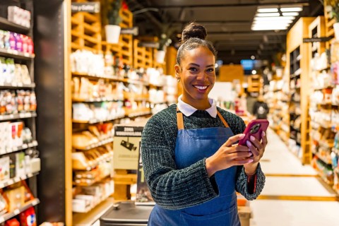 Smiling saleswoman using a smartphone in an organic supermarket with shelves full of natural and