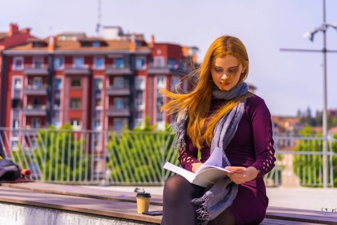 Beautiful woman in purple dress and scarf reading a book in a park in the city, imagining and