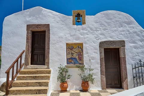 Traditional Greek building with white wall, two doors, bell and staircase under blue sky,