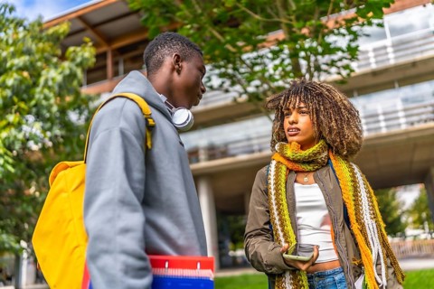 Low angle view photo of two diverse students talking and sharing mobile standing outside the