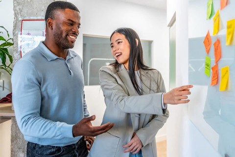 Two smiling businesspeople discussing work related tasks pointing at sticky notes on whiteboard in