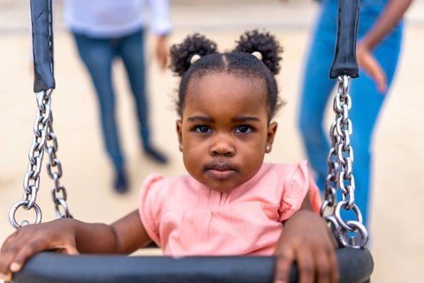Cute african baby girl sitting on swing in a public park next to her family
