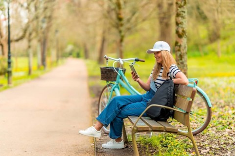 Blonde female tourist wearing casual clothing, using smartphone and resting on park bench with