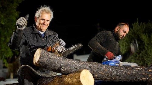 ©Udo Rein, An elderly man gives a thumbs up while sitting next to a woodworker with a chainsaw,